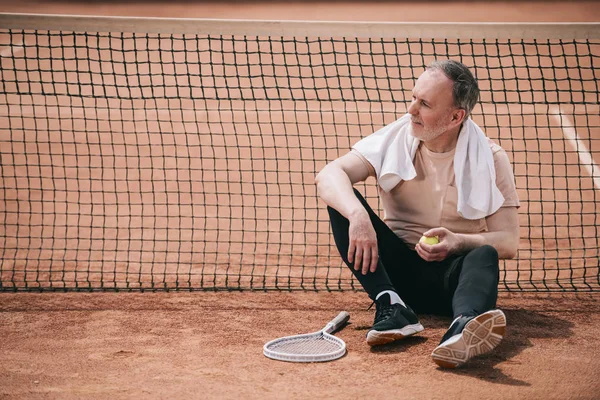 Älterer Mann mit Handtuch und Tennisutensilien ruht neben Netz auf Tennisplatz — Stockfoto