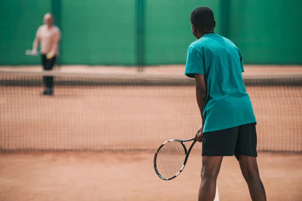 Избирательный фокус пожилого африканского мужчины, играющего в теннис с другом на корте — стоковое фото