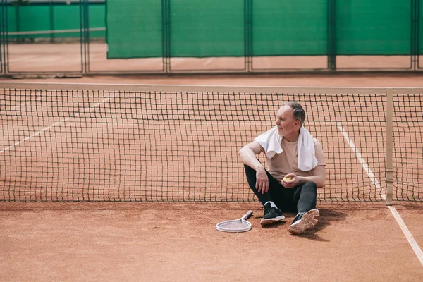 Älterer Mann mit Handtuch und Tennisutensilien ruht neben Netz auf Tennisplatz — Stockfoto