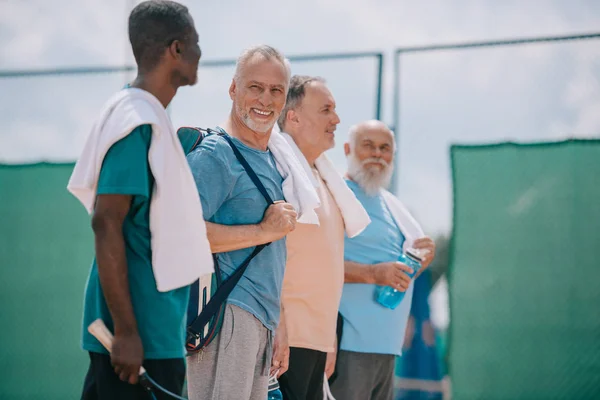 Side view of multicultural elderly men with towels and tennis equipment on tennis court — Stock Photo