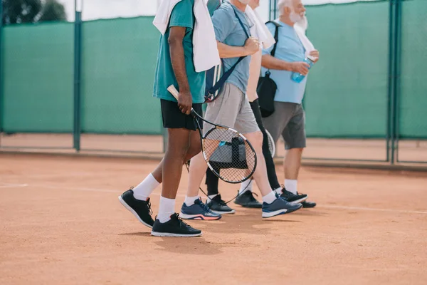 Vue partielle d'hommes âgés multiculturels avec équipement de tennis marchant sur le court — Photo de stock