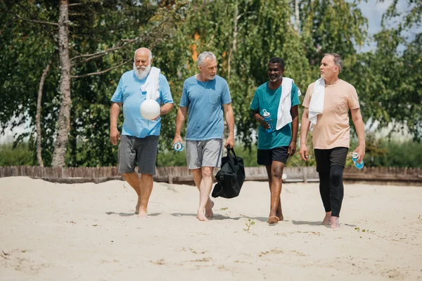 Multiethnische ältere Volleyballspieler mit sportlichen Wasserflaschen, die nach dem Spiel am Strand spazieren gehen — Stockfoto