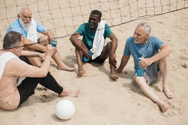 Jogadores de voleibol idosos sorridentes multiétnicos com garrafas de água esportivas descansando após o jogo — Fotografia de Stock