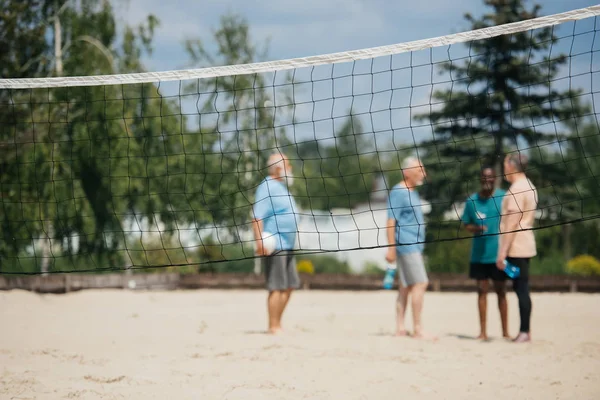 Selective focus of net and interracial old men with volleyball ball on sandy beach — Stock Photo