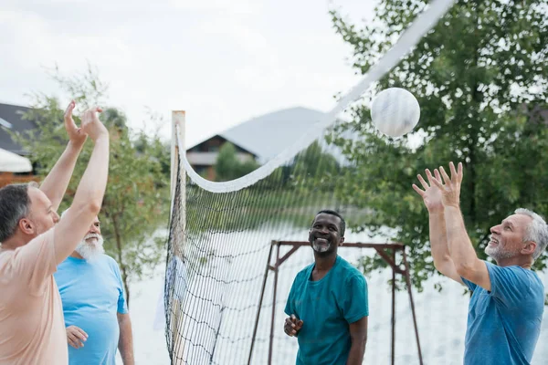 Sonrientes viejos amigos multiculturales jugando voleibol en la playa en el día de verano - foto de stock