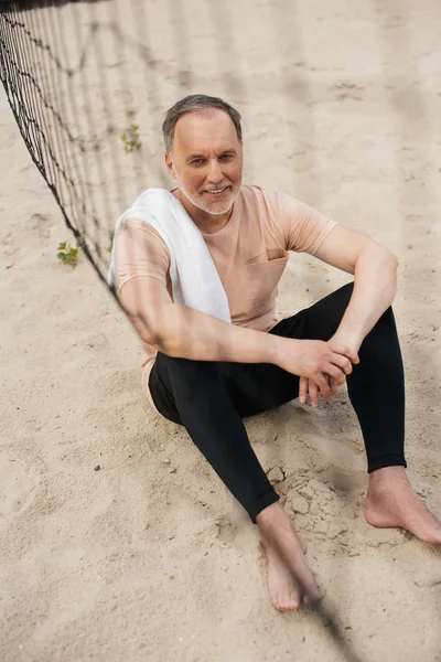 Homme âgé souriant avec serviette reposant près du filet sur la plage de sable après avoir joué au volley-ball — Photo de stock