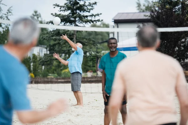 Selective focus of multicultural old friends playing volleyball on beach on summer day — Stock Photo