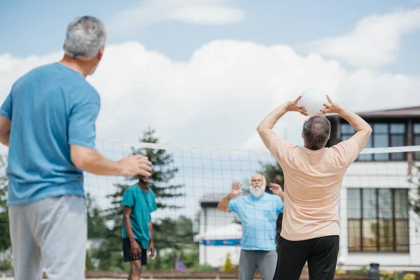 Selective focus of multicultural old friends playing volleyball on beach on summer day — Stock Photo
