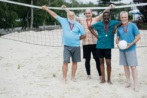 Smiling interracial elderly volleyball players with medals standing on sandy beach — Stock Photo