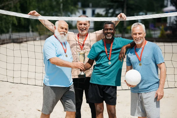 Retrato de amigos ancianos multiétnicos sonrientes con pelota de tenis, medallas y copa de campeones en la playa - foto de stock