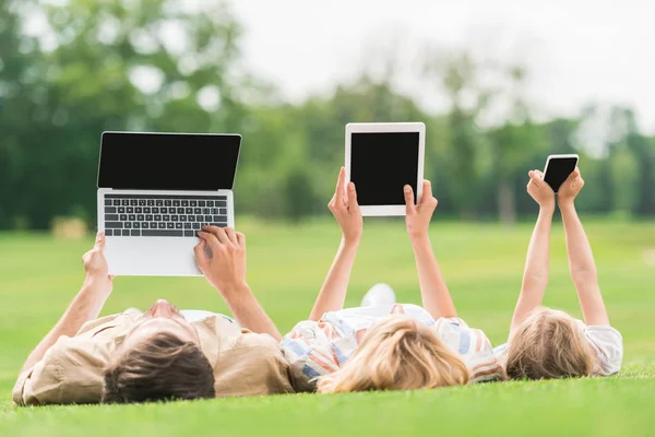 Famiglia sdraiata sull'erba e utilizzando dispositivi digitali con schermi vuoti — Foto stock