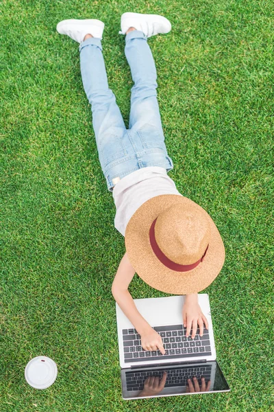 Вид сверху на ребенка в шляпе, лежащего на траве и с помощью ноутбука — стоковое фото