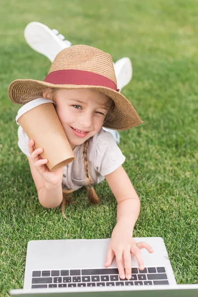 Adorable child holding paper cup and smiling at camera while using laptop on green meadow — Stock Photo
