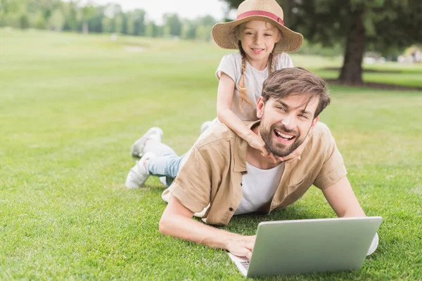 Happy father and daughter using laptop and smiling at camera while lying on grass in park — Stock Photo