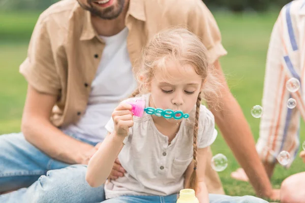 Recortado tiro de padres mirando lindo poco hija soplando pompas de jabón en parque - foto de stock