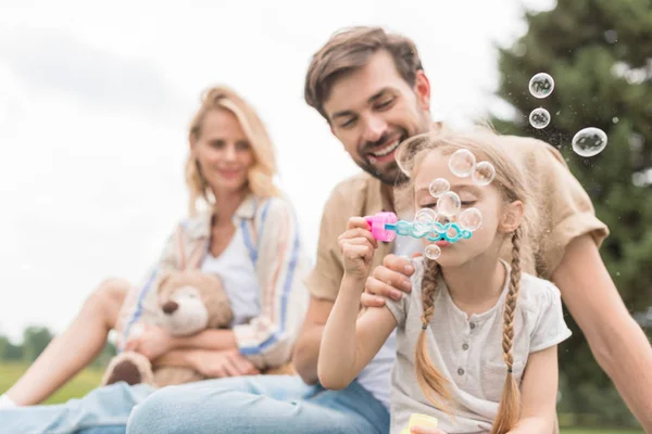 Mother holding teddy bear and looking at father and daughter blowing soap bubbles in park — Stock Photo