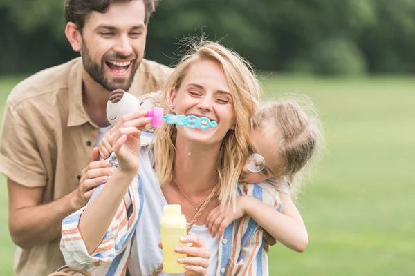 Glückliche Familie mit einem Kind, das sich im Park umarmt und Seifenblasen pustet — Stockfoto