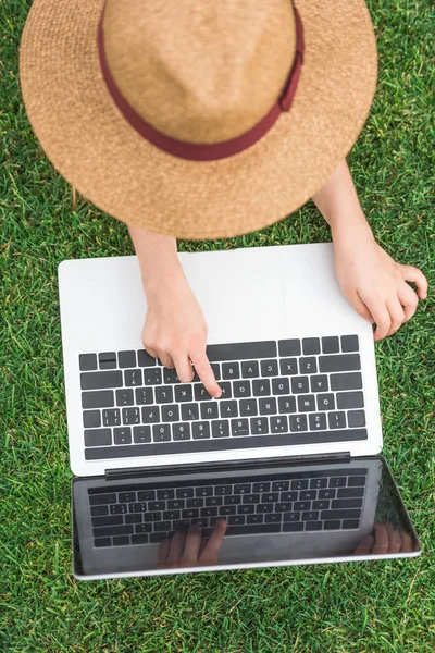 Вид сверху на ребенка в шляпе с помощью ноутбука на зеленой траве — стоковое фото