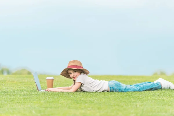 Adorable enfant en chapeau souriant à la caméra tout en utilisant un ordinateur portable sur prairie verte — Photo de stock