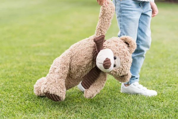 Plan recadré d'un enfant tenant un ours en peluche debout sur une pelouse verte — Photo de stock