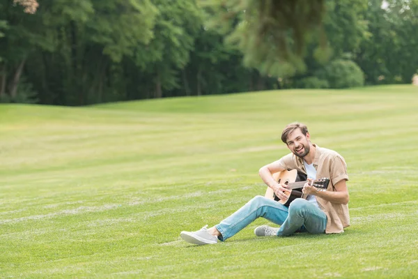 Joven feliz sentado en prado verde y tocando la guitarra acústica - foto de stock