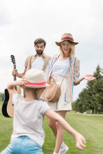 Hermosa madre y feliz padre con la guitarra acústica mirando linda hija divirtiéndose en el parque - foto de stock