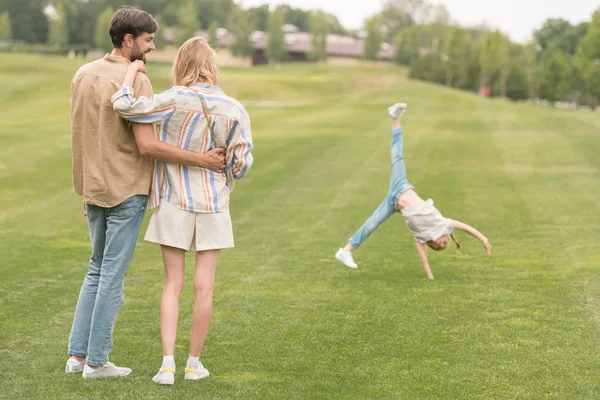 Счастливые родители обнимаются и смотрят на милую маленькую дочь, играющую на руках в парке — стоковое фото