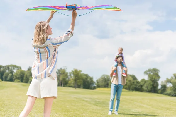 Frau hält bunten Drachen, während Vater kleine Tochter im Park am Hals trägt — Stockfoto