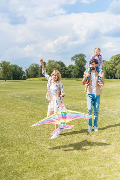 Счастливые молодые родители и милая маленькая дочка, играющая с разноцветным змеем в парке — стоковое фото
