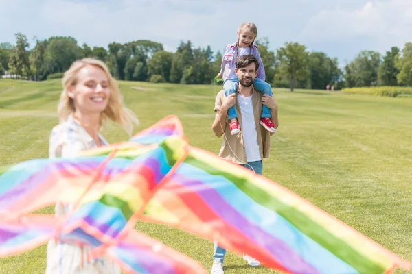 Heureux père portant petite fille sur le cou tandis que la mère tenant cerf-volant coloré dans le parc — Photo de stock