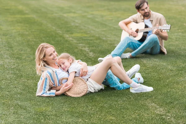 Glückliche Mutter und Tochter umarmen sich auf dem Rasen, während der Vater hinter Gittern spielt — Stockfoto