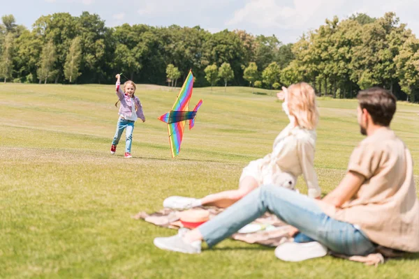 Eltern sitzen auf Plaid und schauen Tochter beim Spielen mit buntem Drachen im Park zu — Stockfoto