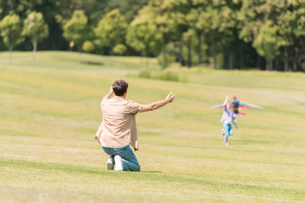 Vista trasera del padre con los brazos abiertos mirando a la pequeña hija jugando con cometa en el parque - foto de stock