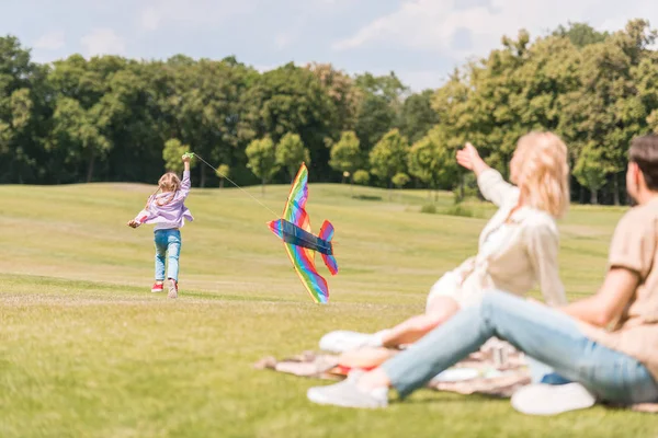 Родители смотрят на дочь, играющую с цветным змеем в парке — стоковое фото