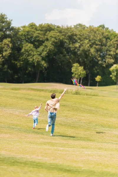 Vista posterior de padre e hija corriendo en el prado y jugando con cometa - foto de stock
