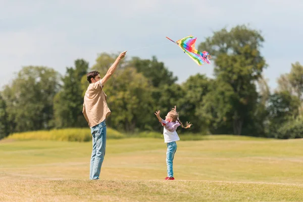 Vista lateral do pai feliz e da filha pequena que joga com o papagaio colorido no parque — Fotografia de Stock