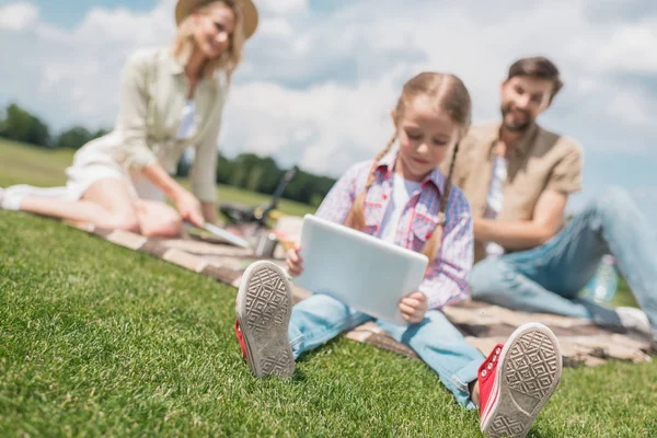 Niño usando tableta digital mientras los padres se sientan detrás en el picnic - foto de stock