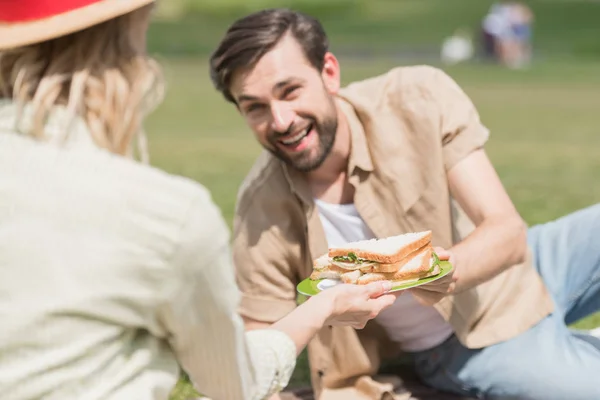Young couple holding sandwiches while spending time together at picnic in park — Stock Photo