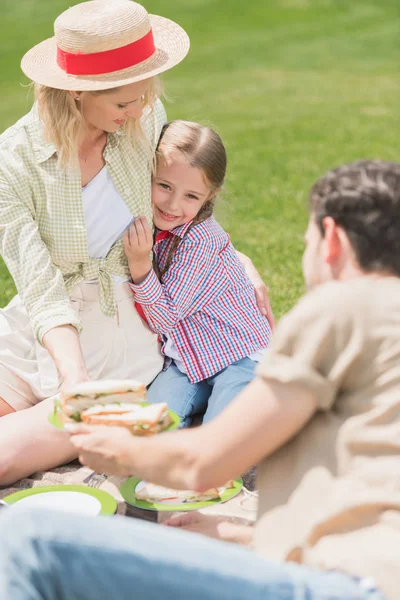 Happy mother and daughter hugging while father holding sandwiches at picnic — Stock Photo