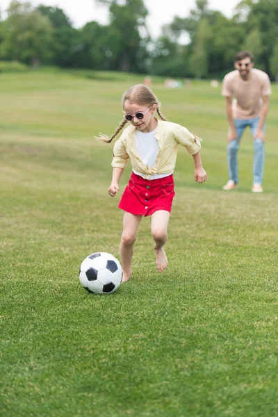 Adorable child in sunglasses playing with soccer ball while father standing behind in oark — Stock Photo