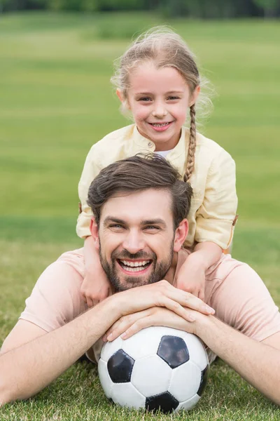 Feliz padre e hija sonriendo a la cámara mientras yacen con pelota de fútbol en la hierba - foto de stock