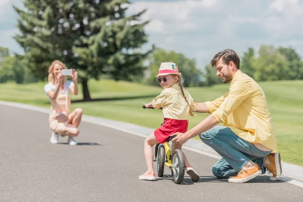 Woman with smartphone photographing father teaching daughter riding bicycle in park — Stock Photo
