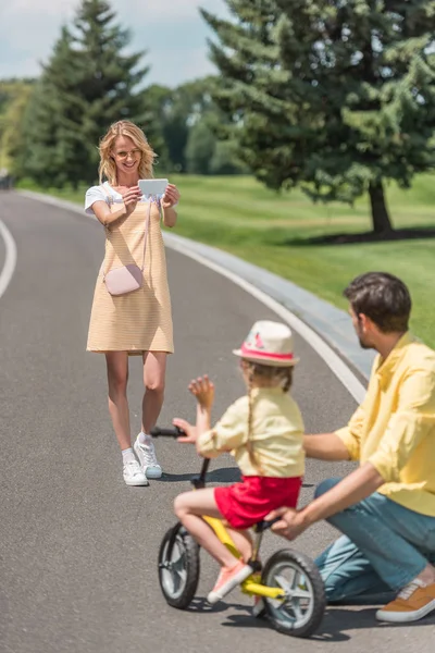 Heureuse jeune femme avec smartphone photographier père enseigner fille équitation vélo dans le parc — Photo de stock