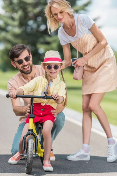 Happy parents looking at cute little daughter riding bicycle in park — Stock Photo