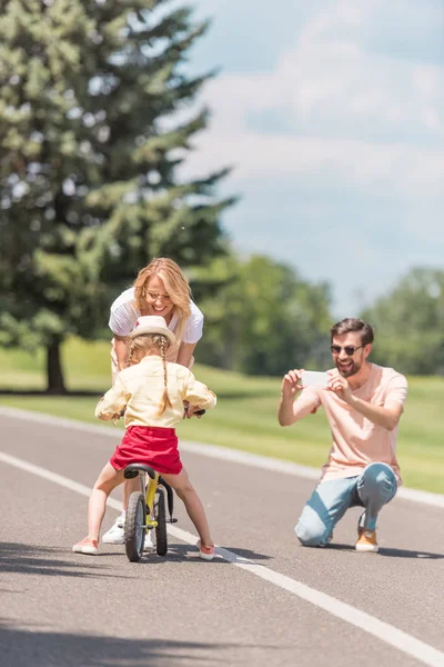 Heureux jeune homme avec smartphone photographier mère souriante enseigner fille équitation vélo dans le parc — Photo de stock