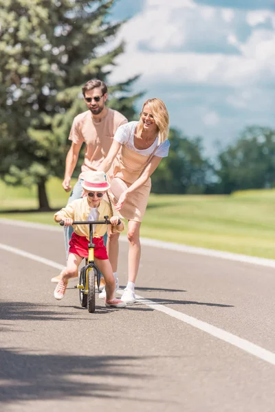 Счастливые молодые родители смотрят на маленькую дочь на велосипеде в парке — стоковое фото