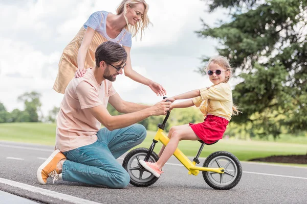 Щасливі батьки дивляться на маленьку дочку верхи на велосипеді в парку — стокове фото