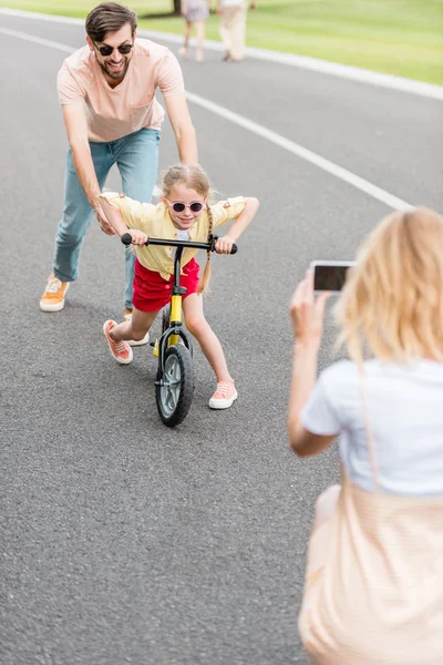 Woman with smartphone photographing happy father and daughter riding bicycle in park — Stock Photo
