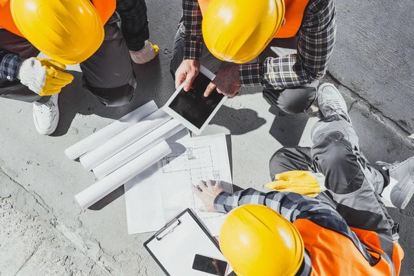 Blick von oben auf drei Bauarbeiter, die auf Beton sitzen und Baupläne diskutieren — Stockfoto