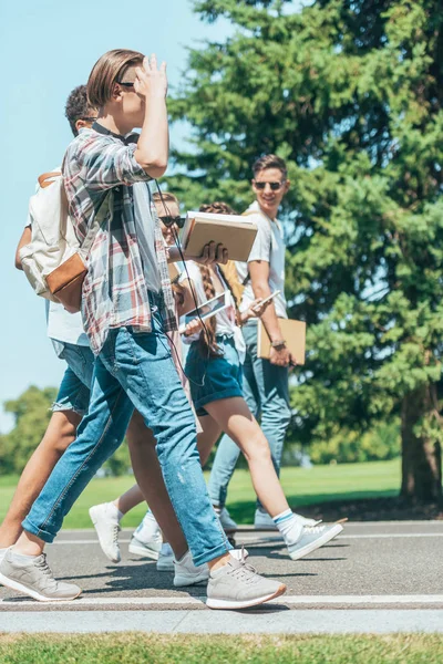 Side view of teenage students with books and gadgets walking together in park — Stock Photo
